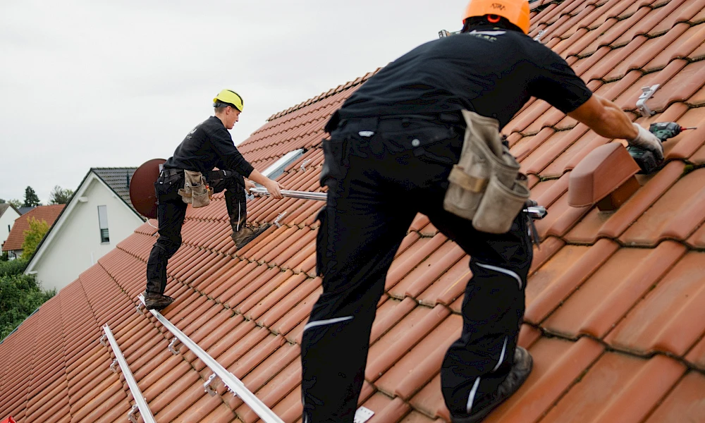Zwei Handwerker montieren Montageschienen für Solarmodule auf einem roten Ziegeldach.